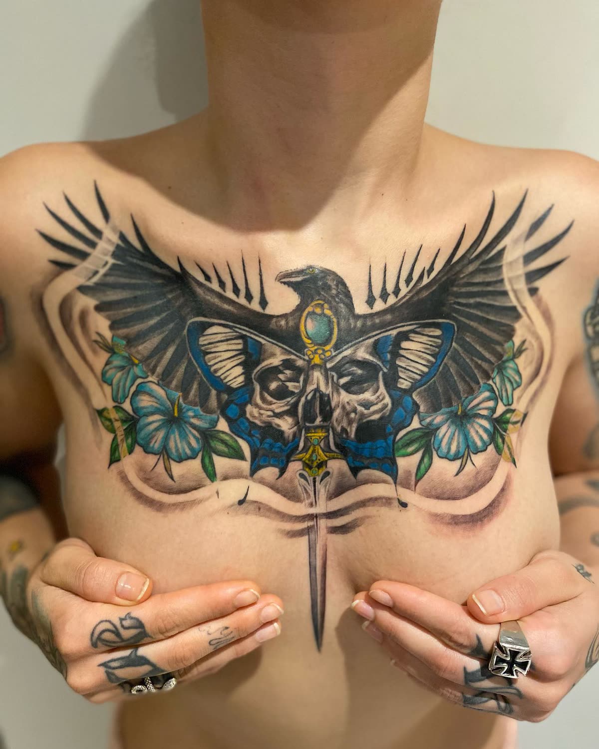 Pecho a color: cuervo con las alas abiertas sobre calaveras, mariposa azul y flores
