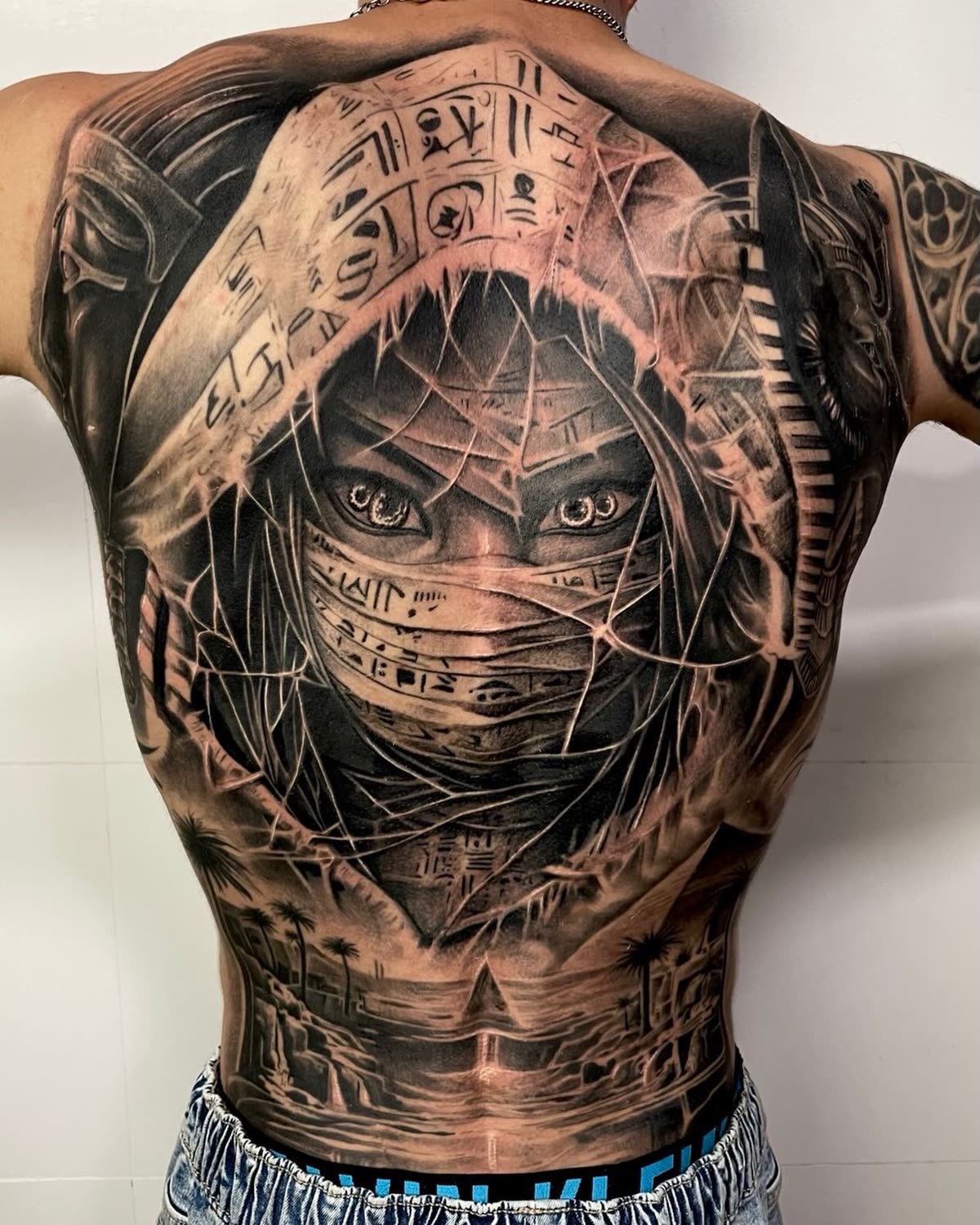 Espalda completa en negro y gris: rostro velado con jeroglíficos, pirámides y palmeras al pie
