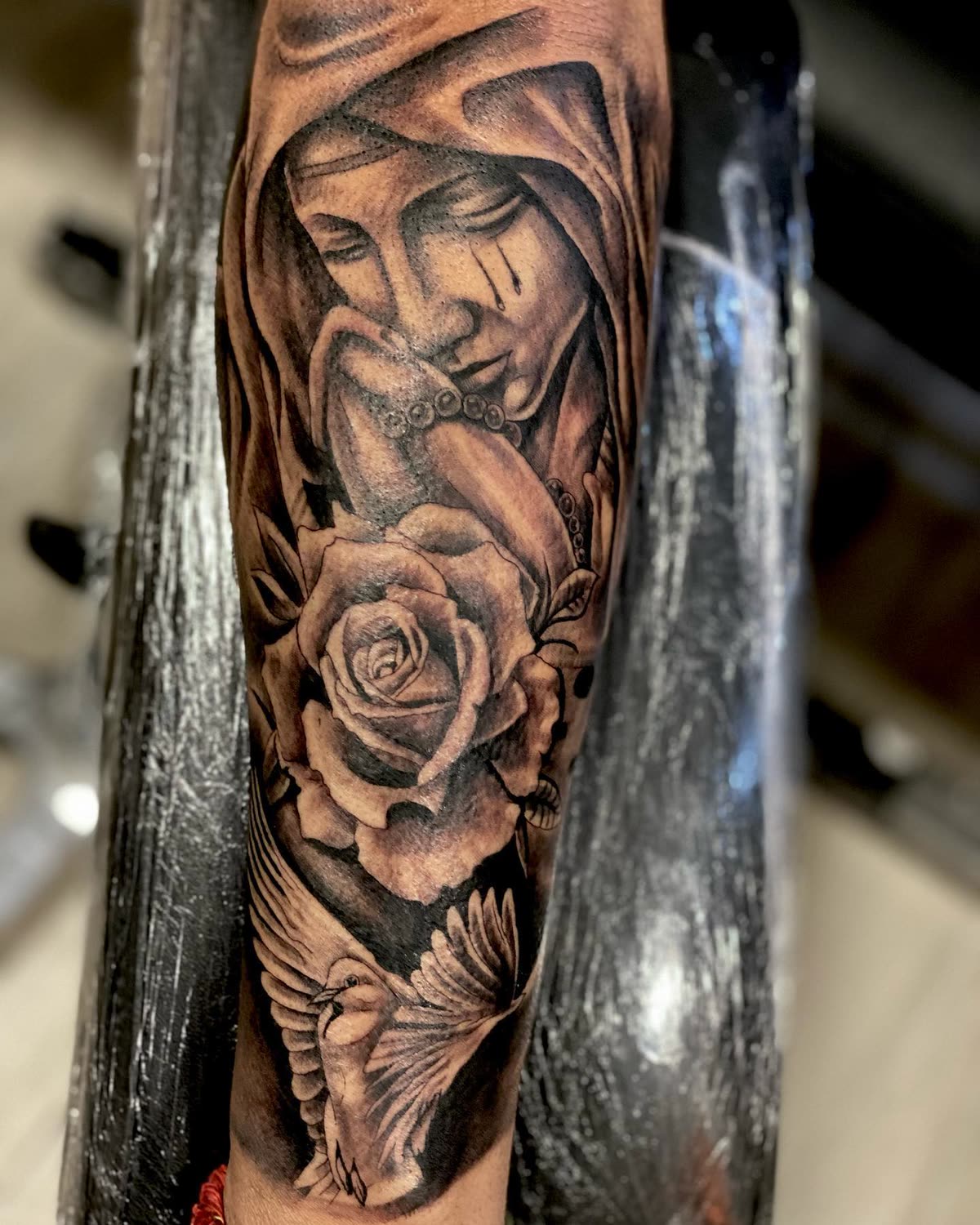 Antebrazo en negro y gris: virgen con rosario, rosa y paloma